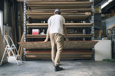 Rear view of man arranging wooden plank in shelf at workshop