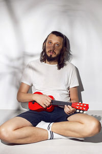 Portrait of a hipster man with a beard sitting against the wall with a small red guitar