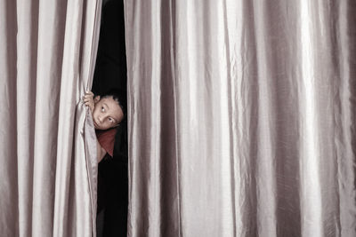 Portrait of young woman hiding behind curtain at home