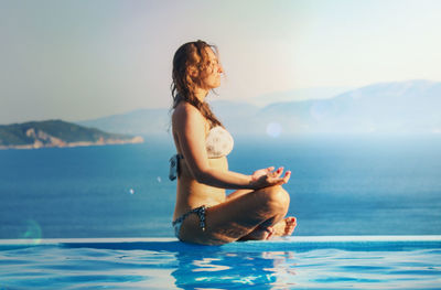 Young woman sitting at infinity pool by sea against sky