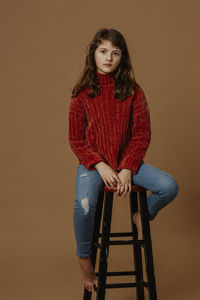 Portrait of serious girl sitting on stool against brown background