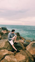 Man sitting on rock at sea shore against sky