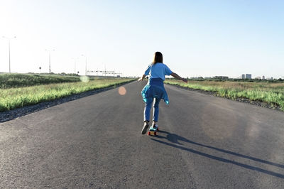 Skater caucasian young woman from back in blue clothes skateboarding on road in summer, active