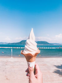 Hand holding ice cream cone at beach against sky