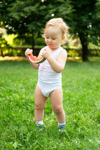 Funny smiling kid boy in white bodysuit eating watermelon at green lawn