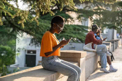 Pleased optimistic african american woman with smartphone outdoors sitting in park in sunny weather