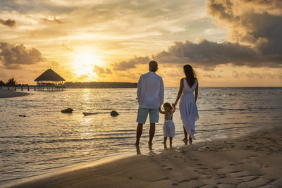 Rear view of couple walking at beach against sky during sunset