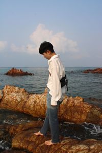 Side view of young man standing on rock by sea against sky