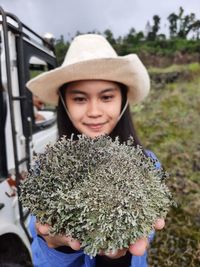 Portrait of smiling young woman holding plant standing by car outdoors