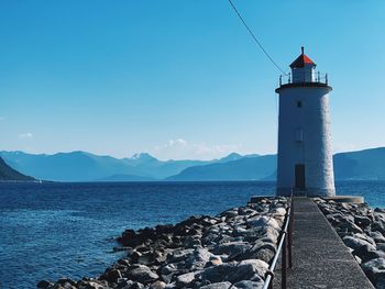Lighthouse by sea and mountain range against clear blue sky, nordic landscape 