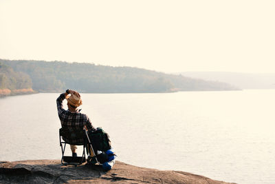 Rear view of man looking at view while sitting on rock by lake