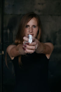 Portrait of young woman aiming gun while standing against wall