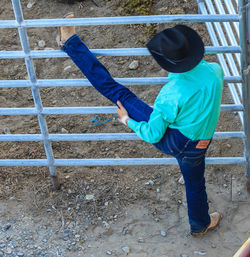 Rear view of cowboy stretching legs on fence at field