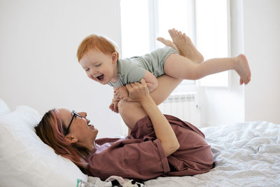 Cheerful mother picking baby boy while lying on bed in bedroom