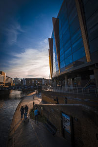People walking by modern city against sky during sunset
