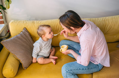 Mother feeding daughter with spoon sitting on sofa at home