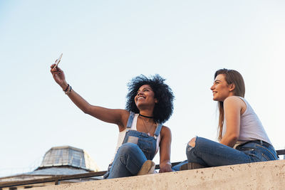 Friends taking selfie on retaining wall against clear sky