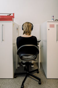 Rear view of female teenage student wearing headphones sitting on chair in library at high school