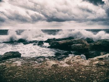 Waves splashing at rocky shore against cloudy sky