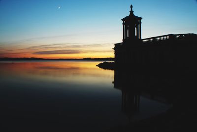 Silhouette of norman tin church by reservoir against sky during sunset