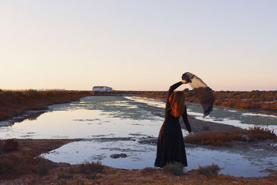 Woman standing by lake against clear sky during sunset