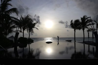 Silhouette palm trees by swimming pool against sky during sunset