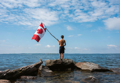 Boy holding canada flag in the air on shore of a lake on a summer day.