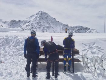 Rear view of skiers standing by wooden bench on snow covered field against mountain