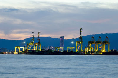 Illuminated factory by sea against sky