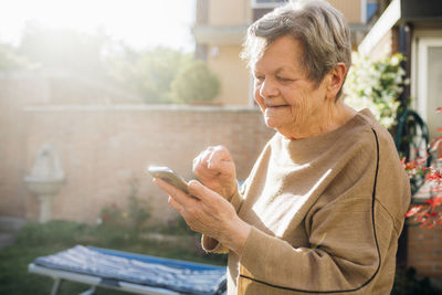 Smiling senior woman using smart phone while standing at backyard during sunny day