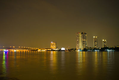 Illuminated buildings by river against clear sky at night