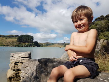 Happy boy sitting on rock by lake against sky