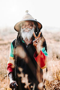 Cheerful old ethnic rastafari with dreadlocks looking at the camera in standing in a dry meadow in the nature