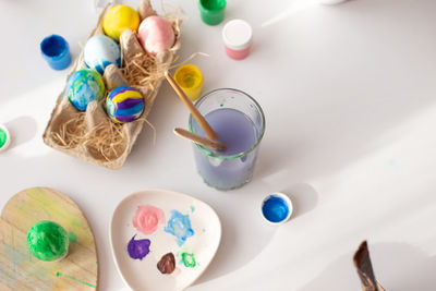 On a white table, in daylight, there are brushes in a glass of water, multi-colored eggs in a tray