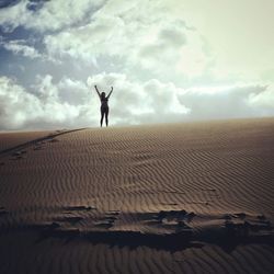 Woman with arms raised standing on sand dune against cloudy sky