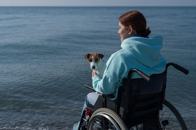 Rear view of woman with dog sitting on wheelchair against sea