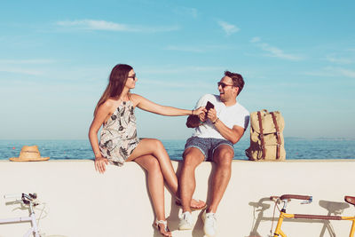 Couple enjoying while sitting on retaining wall by sea against sky