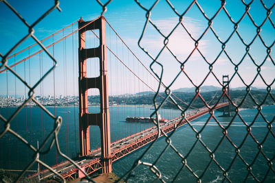 Golden gate bridge over bay against blue sky seen through chainlink fence