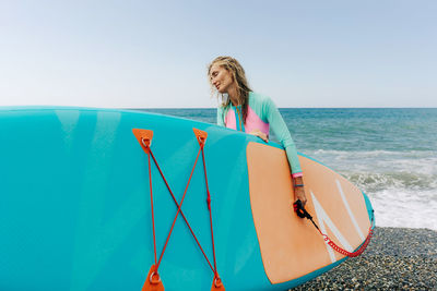 A young athletic blonde woman stands on the beach and holds a paddleboard.