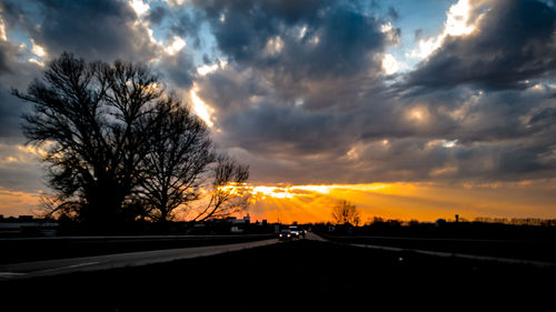 Silhouette trees by road against sky during sunset