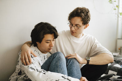 Young man sitting by worried male friend in bedroom