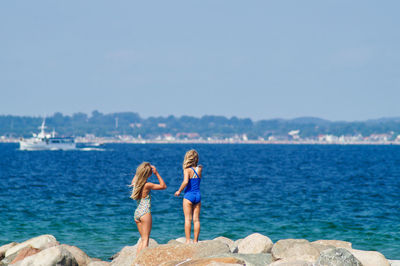 Rear view of women walking on rocks at sea shore against clear sky