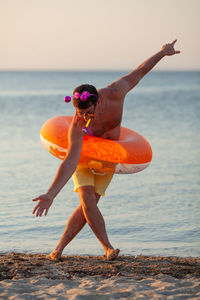 Full length of shirtless man with inflatable ring standing at beach during sunset