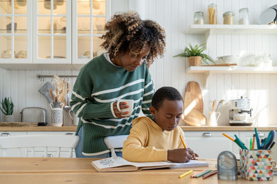 Caring mixed race woman mom with cup look at son handwrite notebook. homeschooling together parent