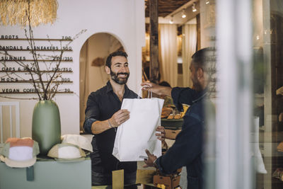 Smiling bearded salesman giving bag to male customer at boutique