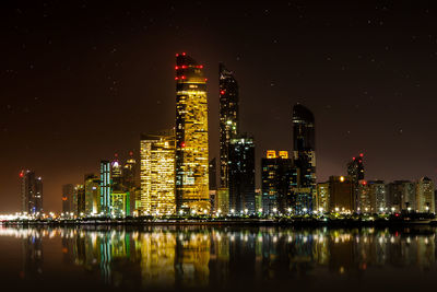 Illuminated buildings by river against sky at night