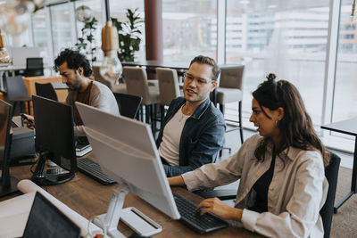 Young male and female colleagues discussing over computer while sitting by businessman at desk in office