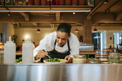 Focused female chef arranging cilantro using tweezers in commercial kitchen