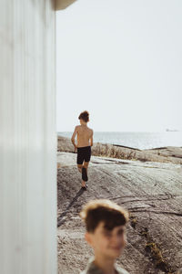 Portrait of teenage boy smiling while shirtless brother walking over rocky land against sky