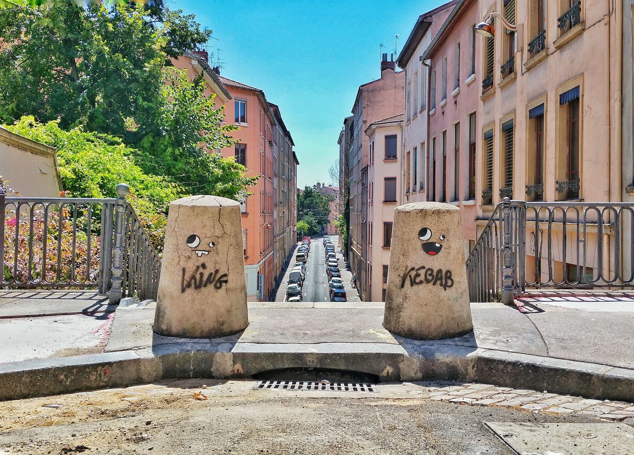 building exterior, architecture, built structure, text, street, tree, city, graffiti, art, sunlight, western script, day, sidewalk, cobblestone, art and craft, outdoors, communication, building, wall - building feature, creativity